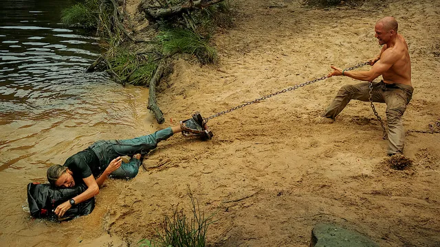 Sasha dragged by a chain shackle on a riverbank while clutching the backpack, confronted by Ben in Apex